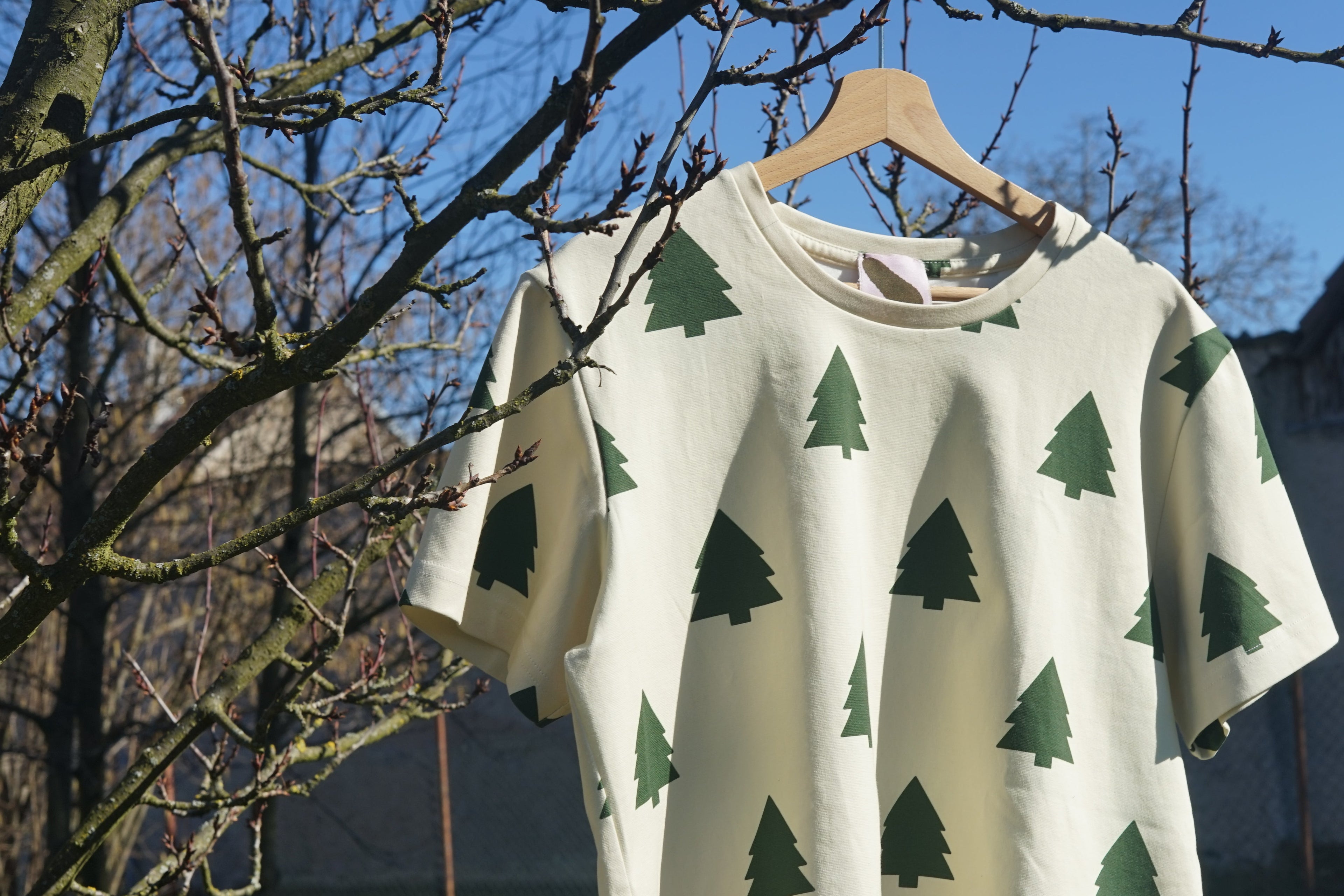 Cream t-shirt with hand-drawn green tree pattern hanging on a wooden hanger in sunlight